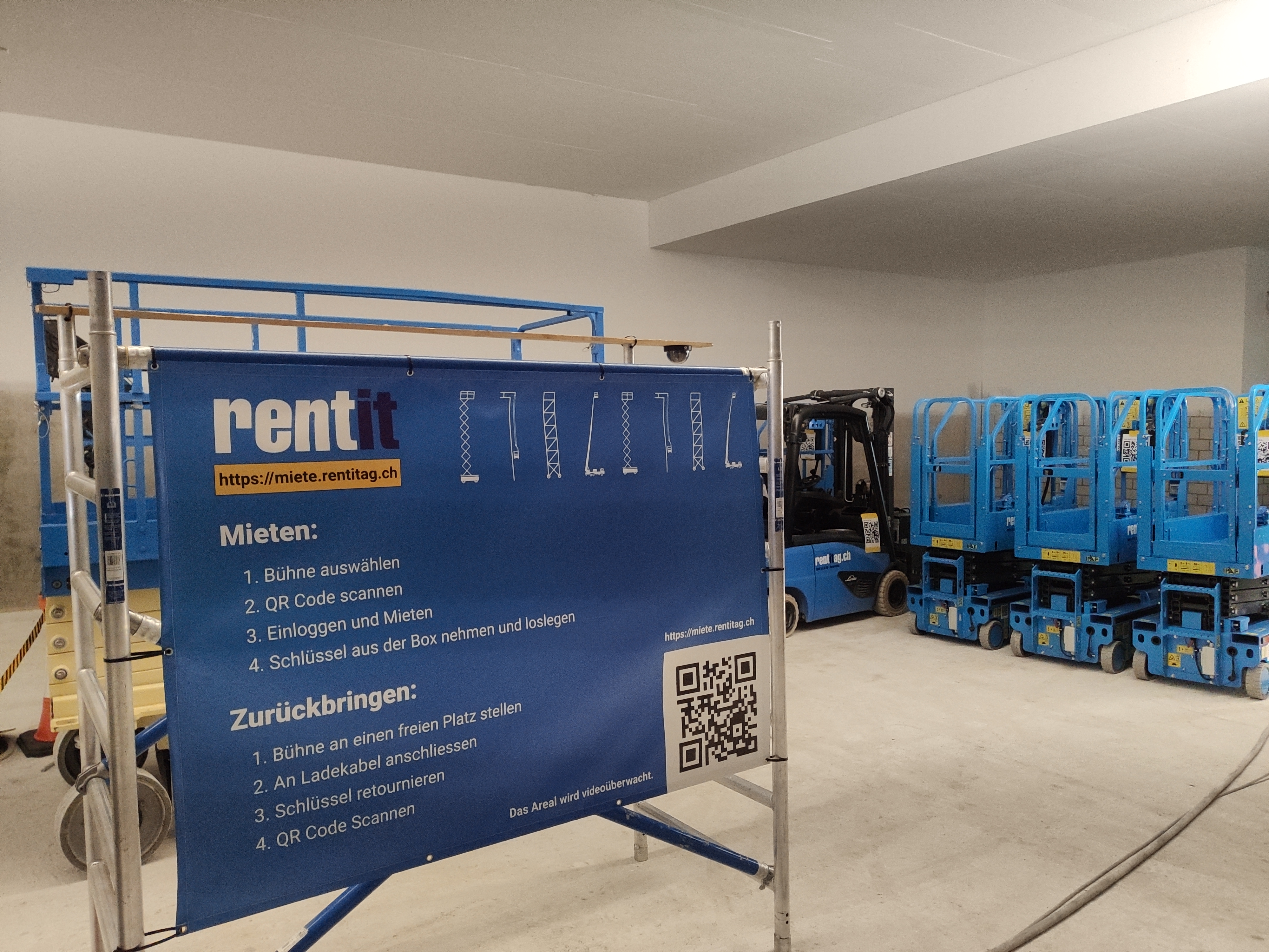 Scherenbühnen und Stapler in einem Kellergeschoss, davor steht ein Plakat mit Anleitungen zur Mietstation