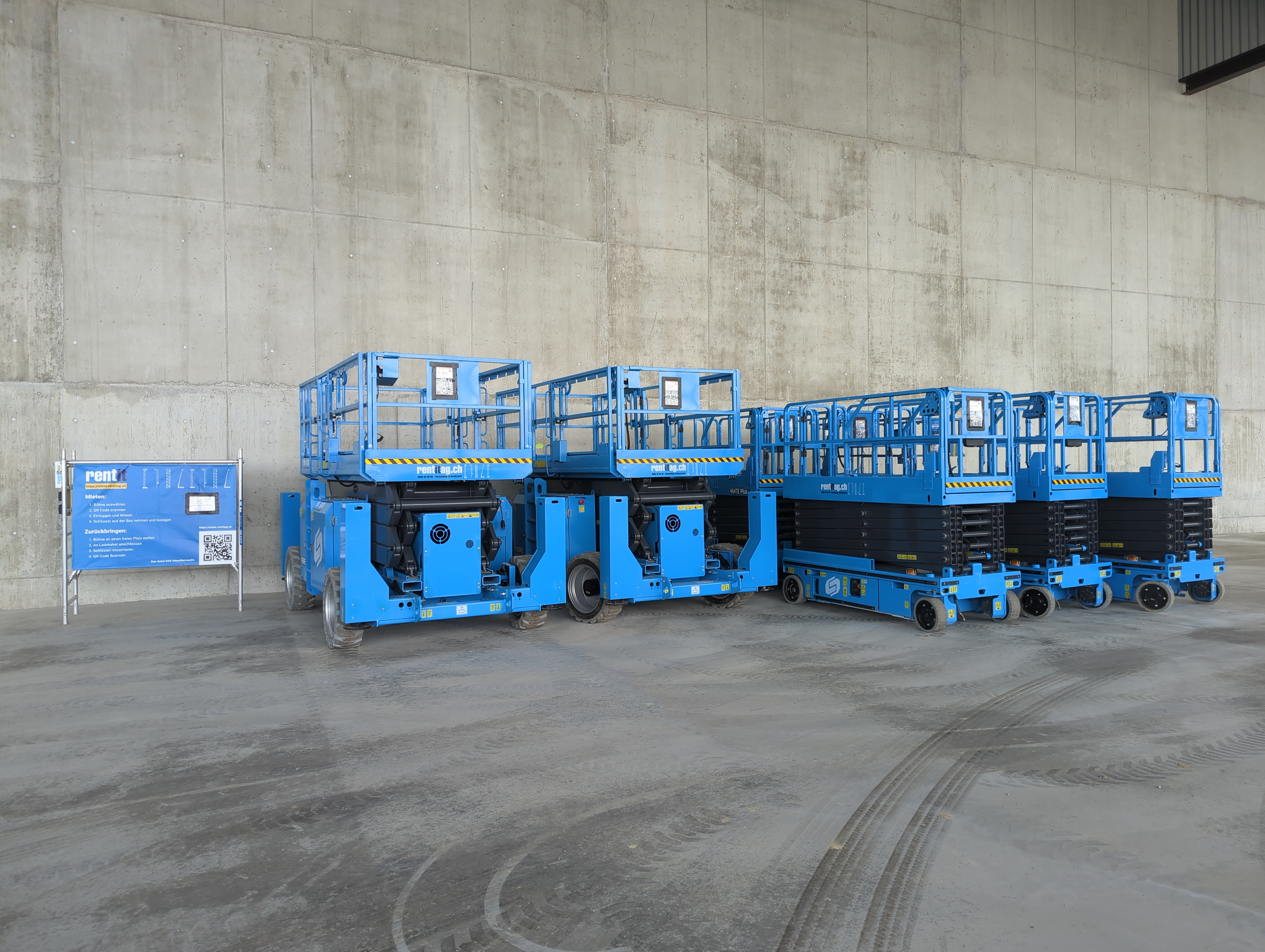 In einer Halle stehen mehrere kleinere und grössere Scherenbühne. Daneben findet sich ein Plakat mit Anleitungen zur Miete der Geräte